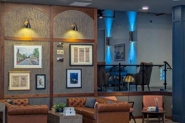 The lobby of Hotel Indigo West Chester featuring a large tiered crystal chandelier, tufted brown leather sofas on a patterned rug, herringbone wood floors, arched wall panels with local artwork and historic photographs, and a raised lounge area with blue accent lighting in the background