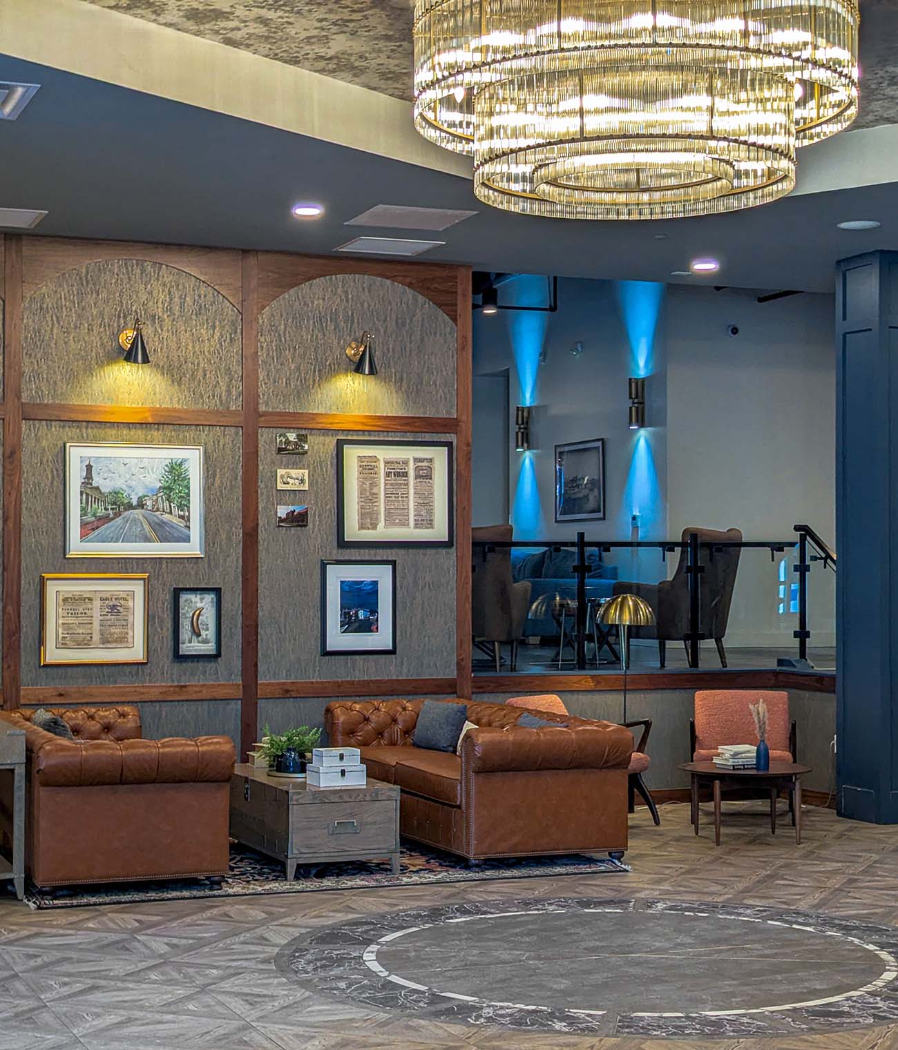 The lobby of Hotel Indigo West Chester featuring a large tiered crystal chandelier, tufted brown leather sofas on a patterned rug, herringbone wood floors, arched wall panels with local artwork and historic photographs, and a raised lounge area with blue accent lighting in the background