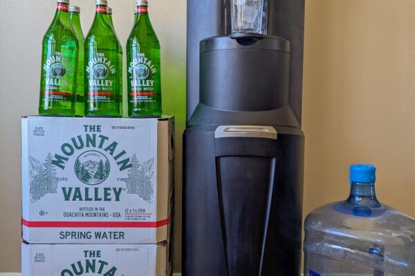 Full Primo bottom-load water dispenser standing tall next to two stacked Mountain Valley spring water cases with three glass bottles on top, and a five-gallon Pure Life jug on the right, in a home setting with a blue abstract painting on the wall.