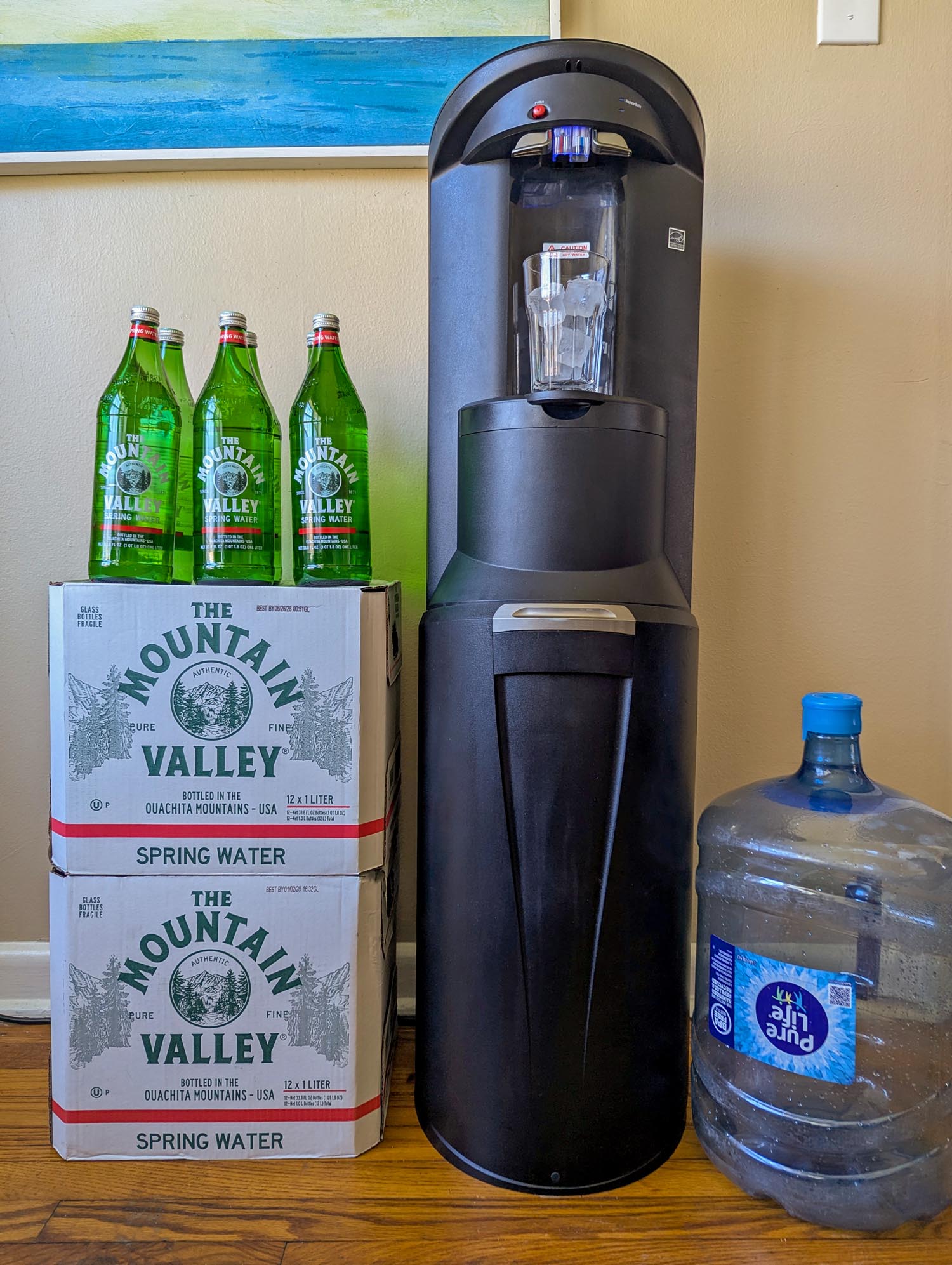 Full Primo bottom-load water dispenser standing tall next to two stacked Mountain Valley spring water cases with three glass bottles on top, and a five-gallon Pure Life jug on the right, in a home setting with a blue abstract painting on the wall.