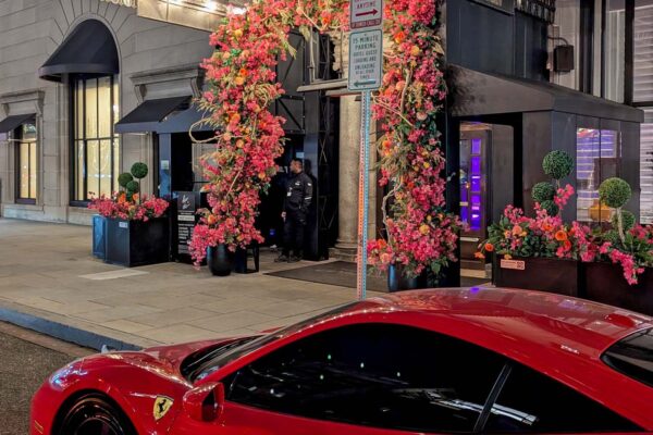 The Hotel Washington DC entrance lit up at night during cherry blossom season, with dramatic pink and orange floral arch installations framing the black entrance canopy displaying the Hotel Washington name in gold lettering, topiary plants flanking the doorway, a doorman standing at the entrance, and the front hood of a red Ferrari visible in the foreground