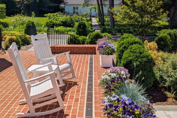 The back brick patio at Brampton 1860 in early spring, featuring two white wooden rocking chairs, a row of white planter boxes overflowing with purple, pink, yellow, and white pansies and violas, neatly trimmed boxwood hedges lining a stone pathway, towering evergreen and deciduous trees