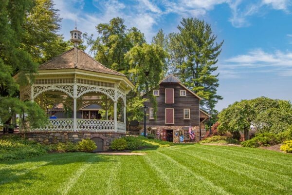 White Victorian gazebo with ornate trim on manicured lawn, surrounded by lush green trees and historic colonial buildings at Peddler