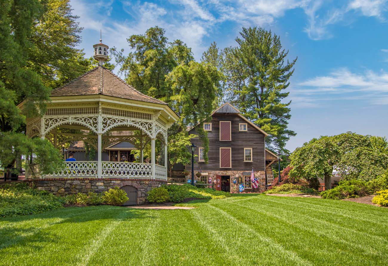 White Victorian gazebo with ornate trim on manicured lawn, surrounded by lush green trees and historic colonial buildings at Peddler