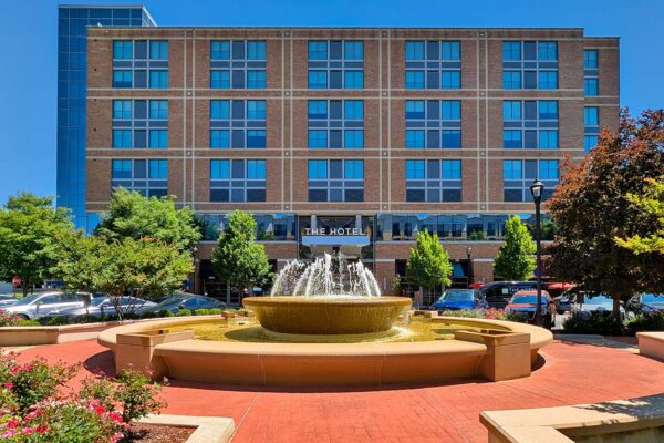 The Hotel at Arundel Preserve exterior on a sunny day, showing the brick and glass facade with a large fountain and red flower beds in the foreground.