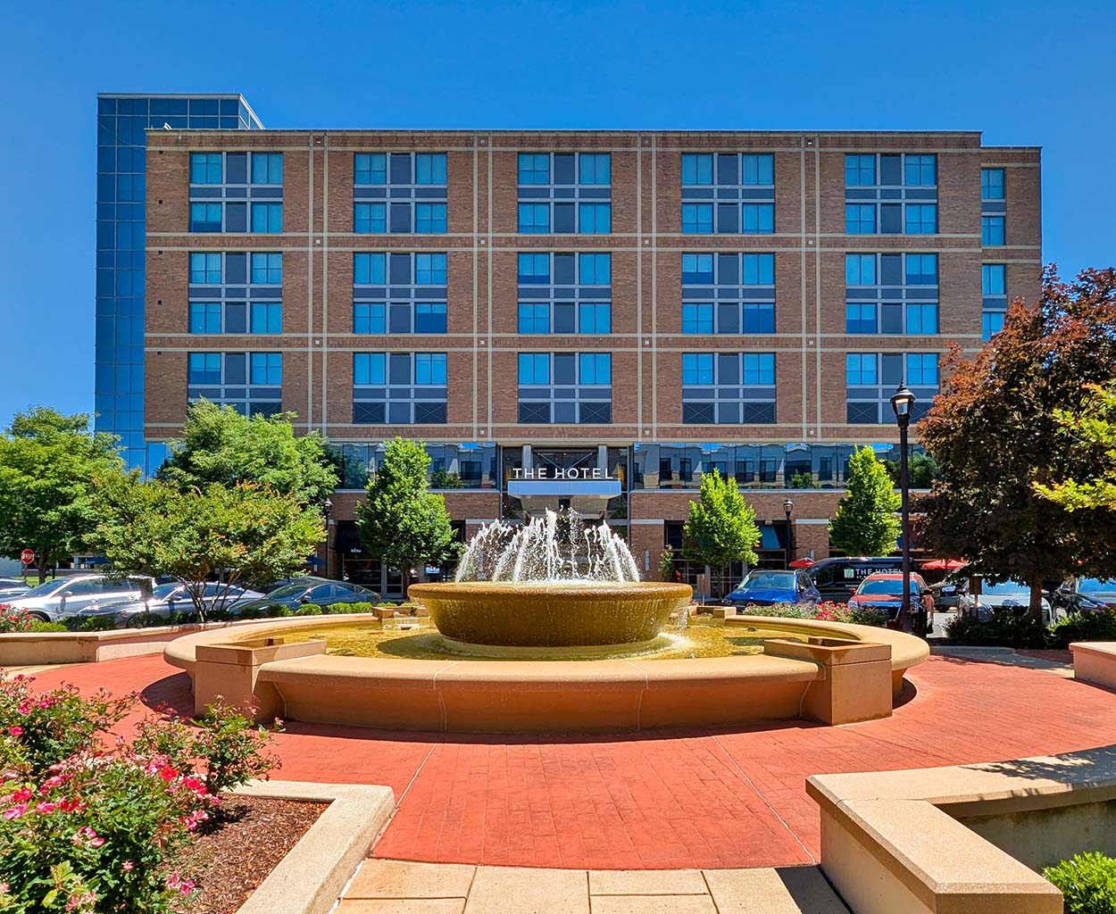 The Hotel at Arundel Preserve exterior on a sunny day, showing the brick and glass facade with a large fountain and red flower beds in the foreground.