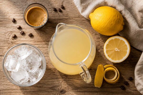 Espresso lemonade ingredients arranged overhead, including a shot of espresso, a pitcher of lemonade, ice, lemon, and lemon peel